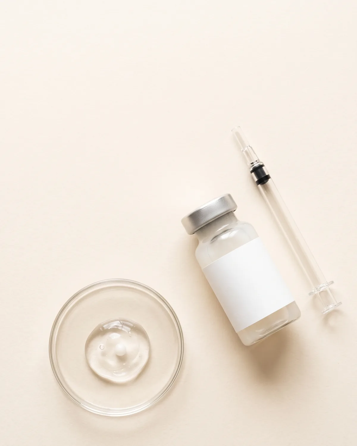 A glass vial with a blank label, a glass mixing applicator and a dish of clear gel on a cream surface